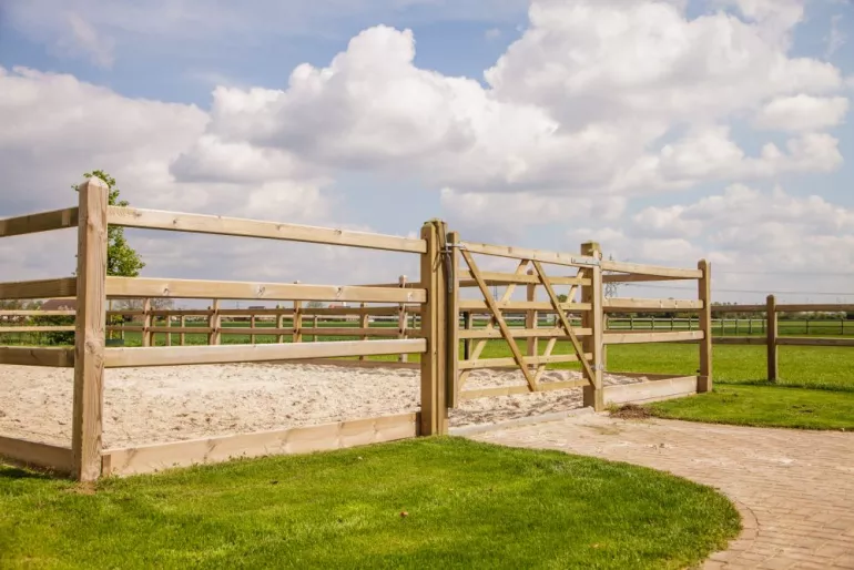 Ein Holztor, das an einem Holzzaun mit quadratischen Pfosten und 3 Stäben befestigt ist, der einen Reitplatz auf einem Feld umschließt